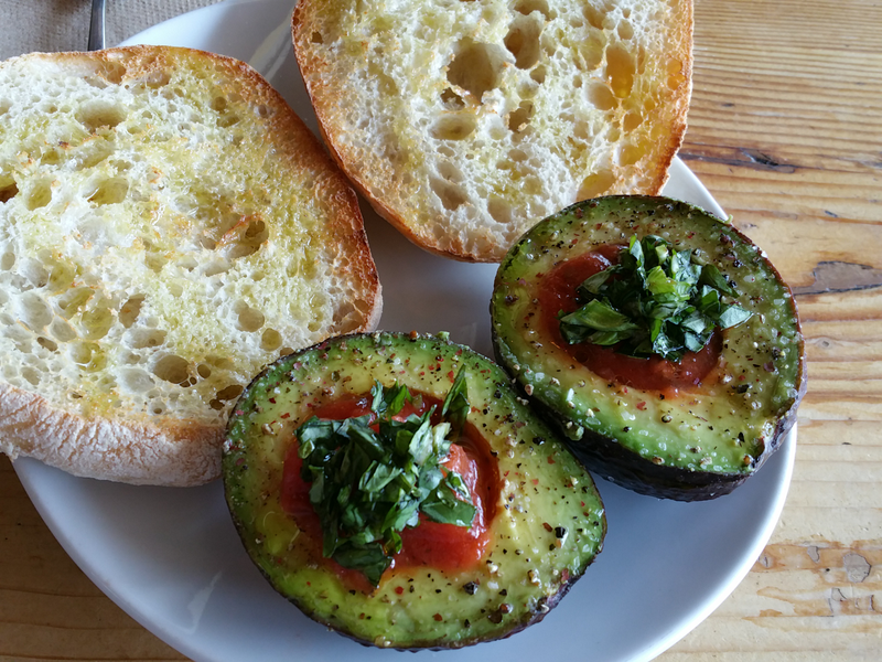 An avocado split with bruschetta, olive oil, fresh basil, salt and pepper. A toasted ciabatta roll drizzled with olive oil to accompany it